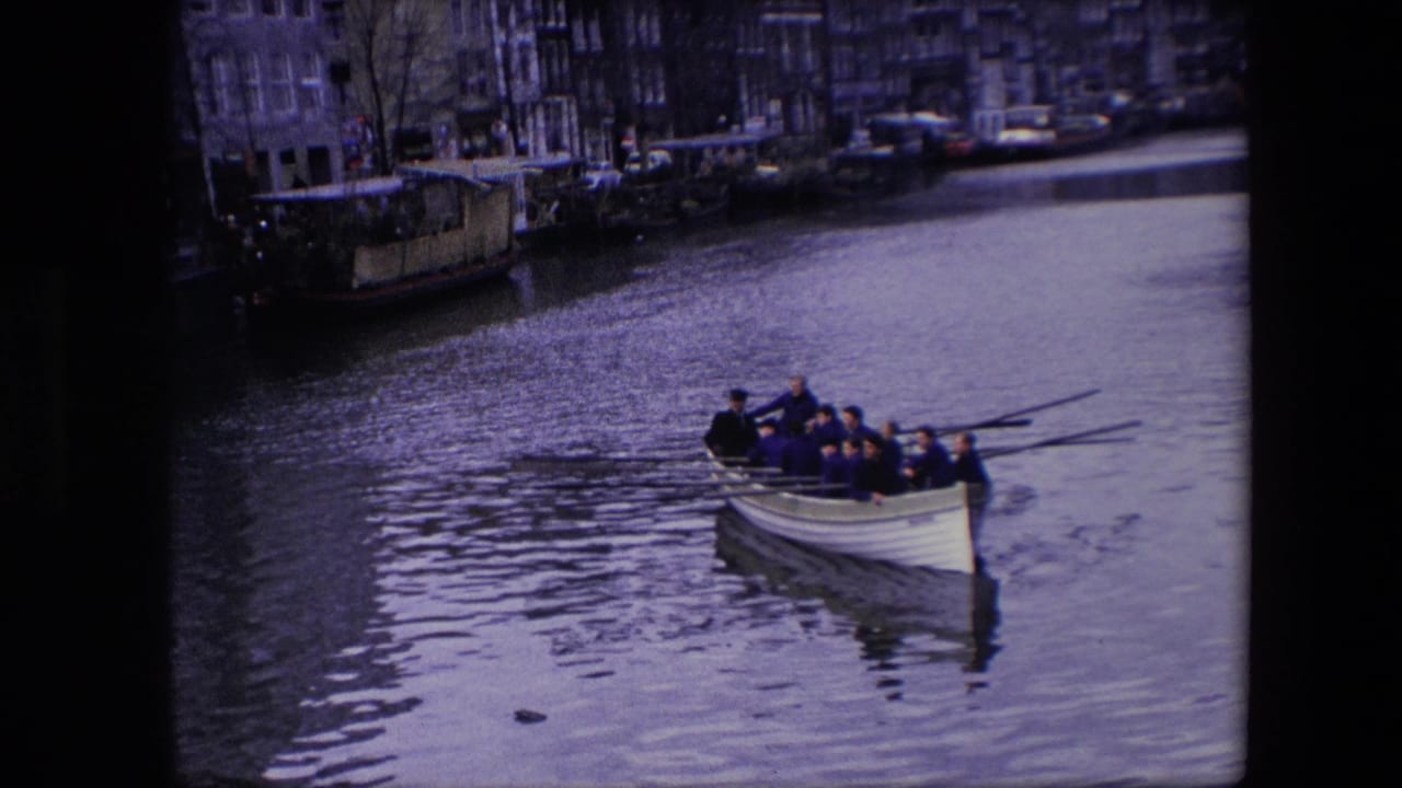 The rowing crew continues along the Amsterdam canal in 1967, with long oars angled over the water and city buildings lining the far bank.