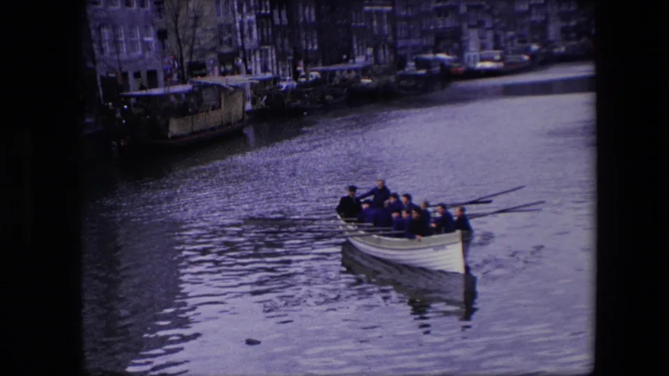 The rowing crew continues along the Amsterdam canal in 1967, with long oars angled over the water and city buildings lining the far bank.