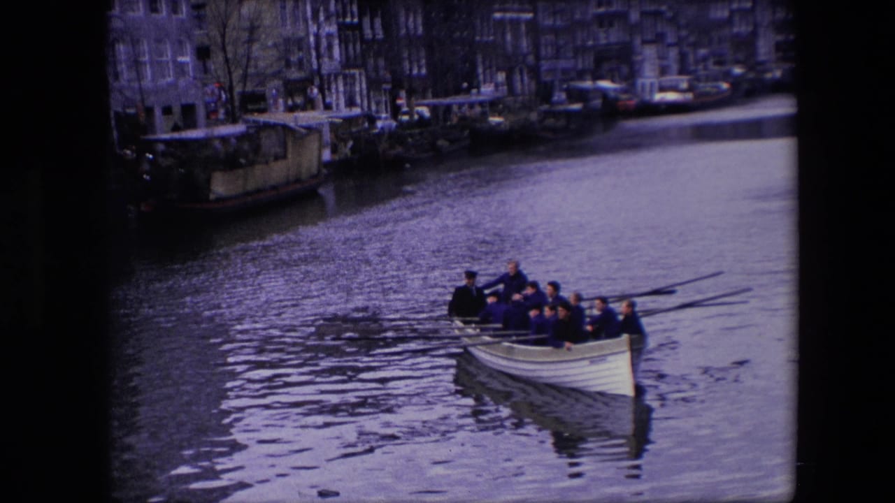 In metadata-identified 1967 Amsterdam footage, a crew of adult men rows a white boat across a canal with buildings and moored vessels in the background.