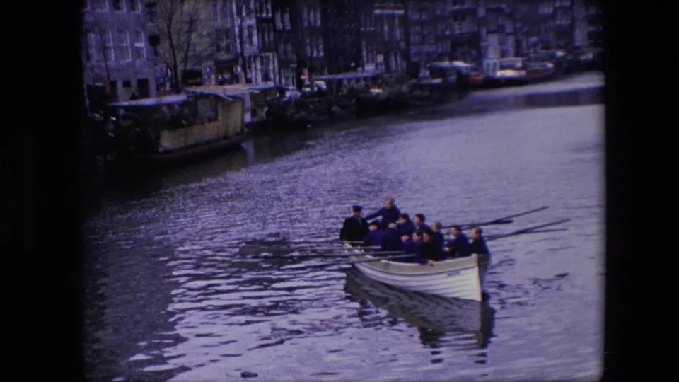 In metadata-identified 1967 Amsterdam footage, a crew of adult men rows a white boat across a canal with buildings and moored vessels in the background.
