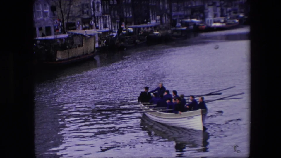 The rowing crew continues along the canal, with several men seated in a white boat and long oars reaching over the water in the Amsterdam footage.
