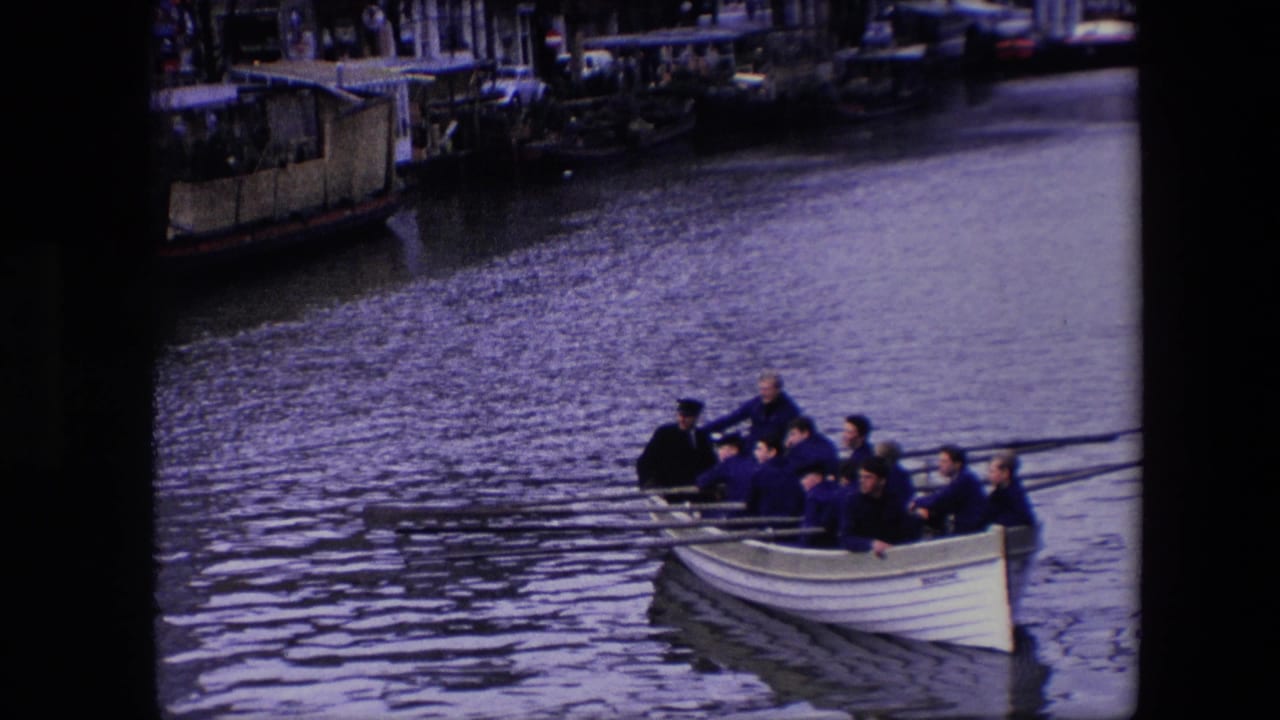 A closer view shows the adult rowing crew in dark jackets and caps as the white boat approaches along the canal, with oars stretched across the water.