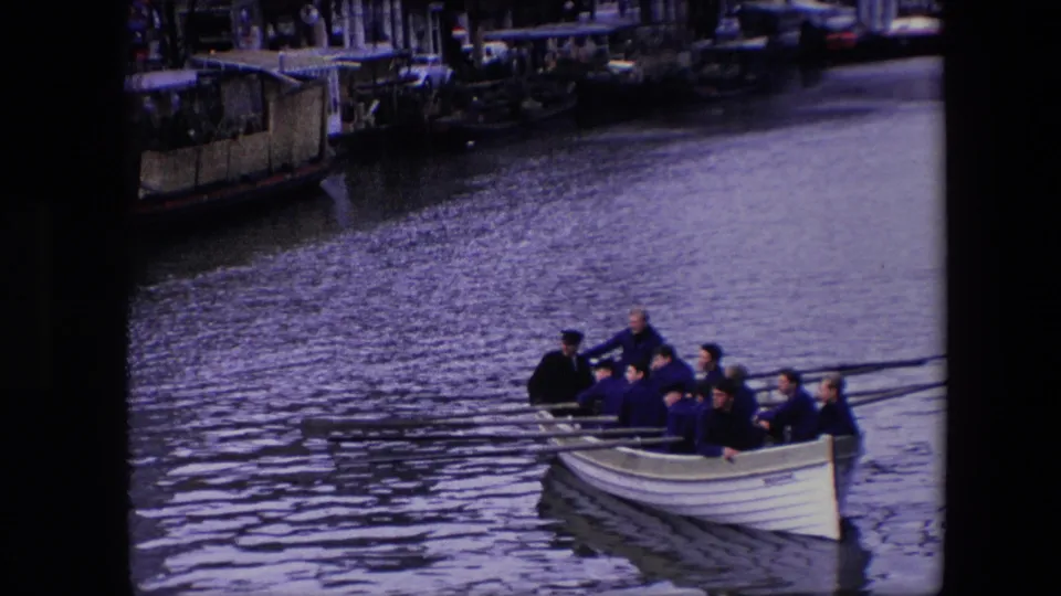 A closer view shows the adult rowing crew in dark jackets and caps as the white boat approaches along the canal, with oars stretched across the water.