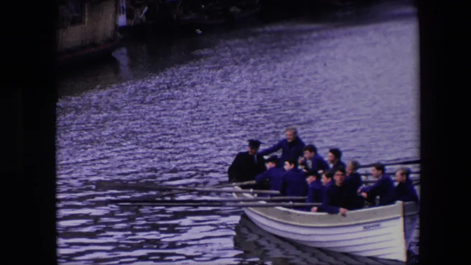 The final frame brings the rowing crew nearest to view, showing multiple adult men in the white boat with long oars and canal water filling most of the frame.