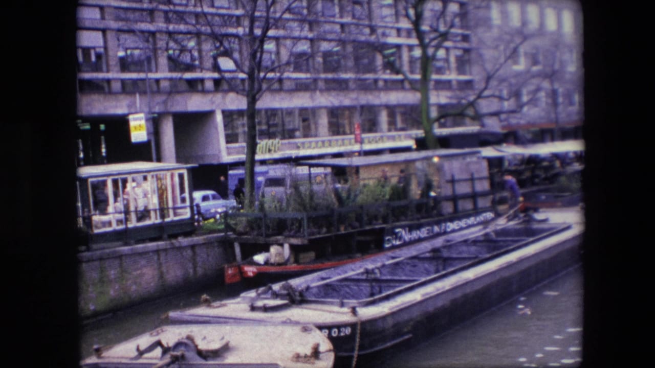 1967 Amsterdam canal scene showing a barge with plants moored along the water, bare trees, parked cars, and multi-story waterfront buildings.