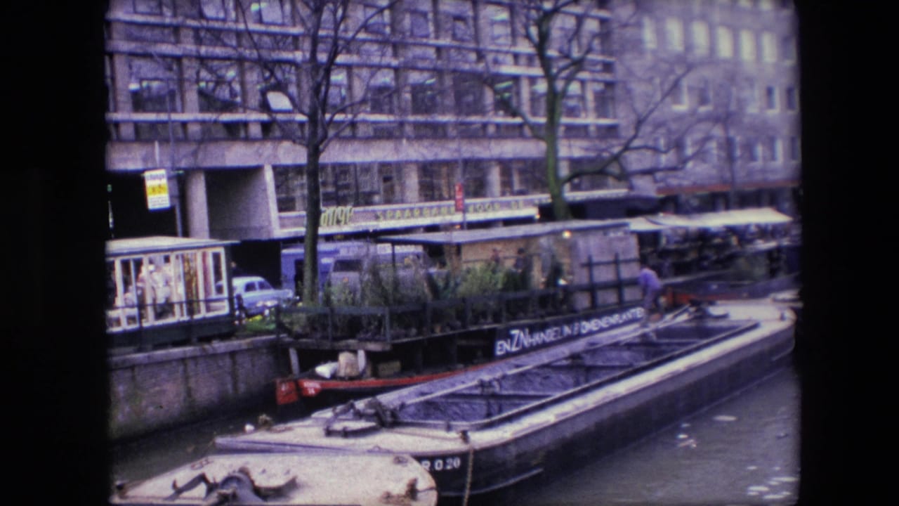 Moored canal barge with plants in front of waterfront buildings and bare trees.