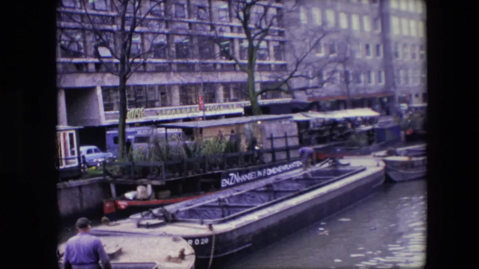 1967 Amsterdam home-movie frame showing a man in a purple shirt walking near the water while a plant-filled canal barge sits beside waterfront buildings.
