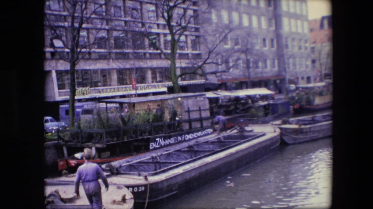 1967 Amsterdam canal footage frame with a man walking along the water's edge beside a barge, bare trees, parked cars, and large waterfront buildings.
