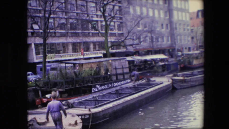 1967 Amsterdam canal footage frame with a man walking along the water's edge beside a barge, bare trees, parked cars, and large waterfront buildings.