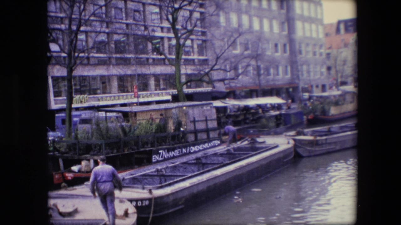 1967 Amsterdam canal scene with a man in a purple sweater walking on a boat, moored barges and houseboats in the foreground, and bare trees and multi-story buildings across the water.