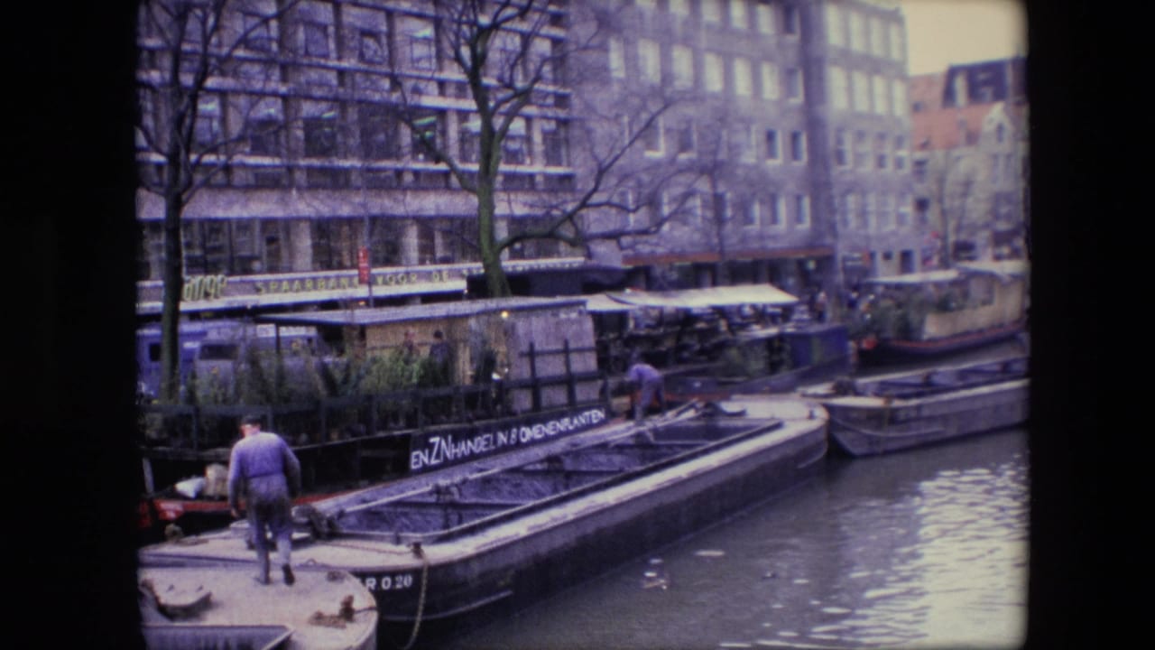 1967 Amsterdam canal view showing a man on the stern of a boat near a moored barge with painted lettering, houseboats, canal water, leafless trees, and waterfront buildings.