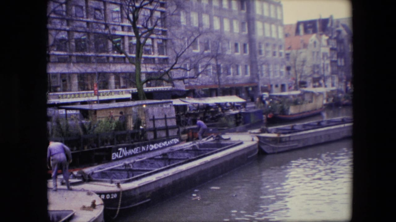 1967 Amsterdam canal footage with a man partly at frame left, a long barge with visible lettering, houseboats, canal water, bare trees, and multi-story buildings behind the quay.