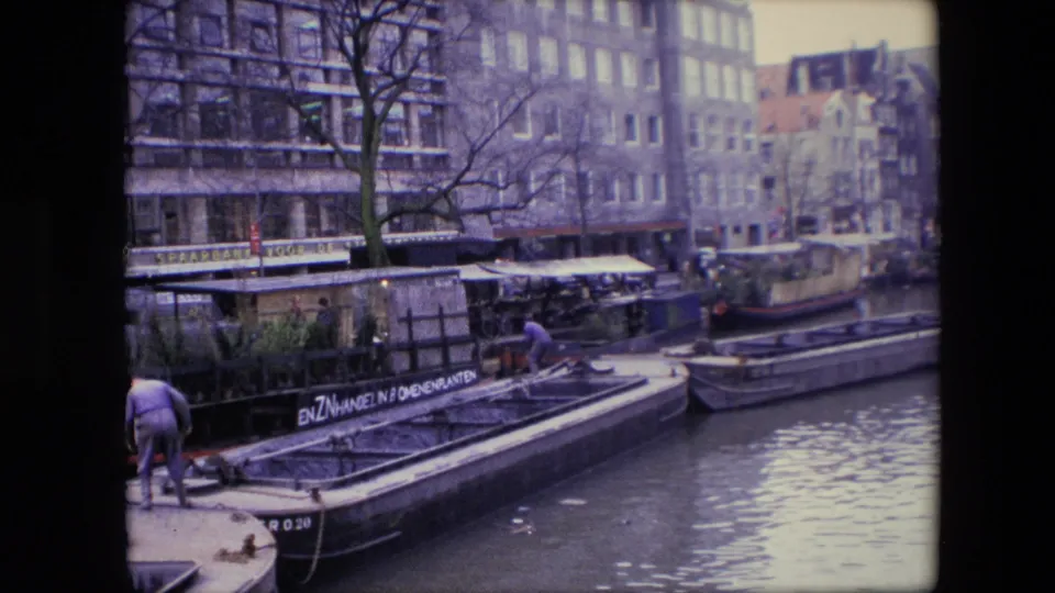 1967 Amsterdam canal footage with a man partly at frame left, a long barge with visible lettering, houseboats, canal water, bare trees, and multi-story buildings behind the quay.