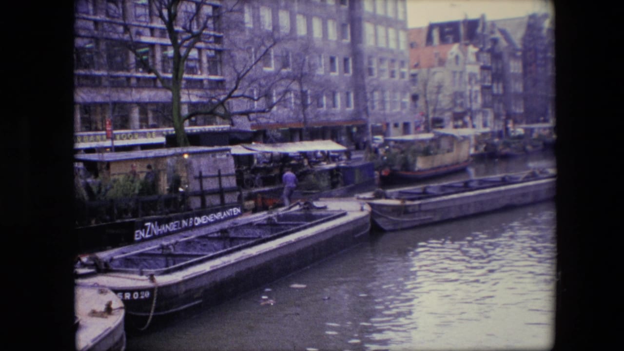 1967 Amsterdam canal view showing a man on a moored boat in the middle distance, a lettered barge, houseboats, rippling water, bare trees, and urban buildings lining the canal.