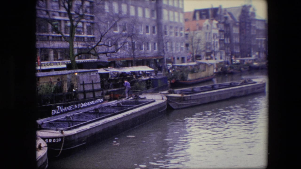 1967 Amsterdam canal scene with flat-bottomed boats, houseboats, older canal buildings, bare branches, rippled water, and a person in blue standing near a dock.