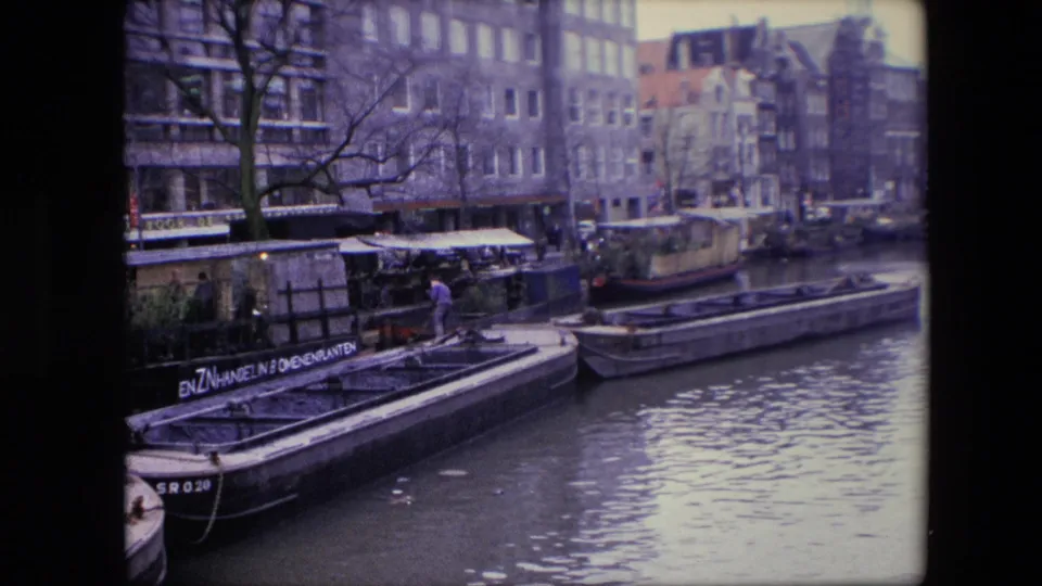 1967 Amsterdam canal scene with flat-bottomed boats, houseboats, older canal buildings, bare branches, rippled water, and a person in blue standing near a dock.