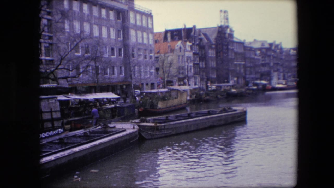 1967 Amsterdam canal footage showing moored houseboats and barges below tall canal-side buildings, with a dockside person partly visible at the left.