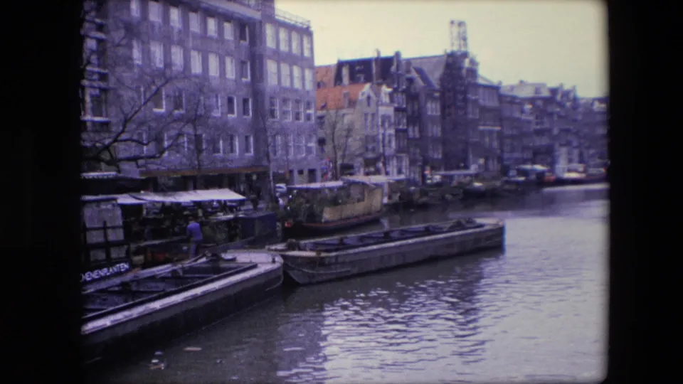 1967 Amsterdam canal footage showing moored houseboats and barges below tall canal-side buildings, with a dockside person partly visible at the left.