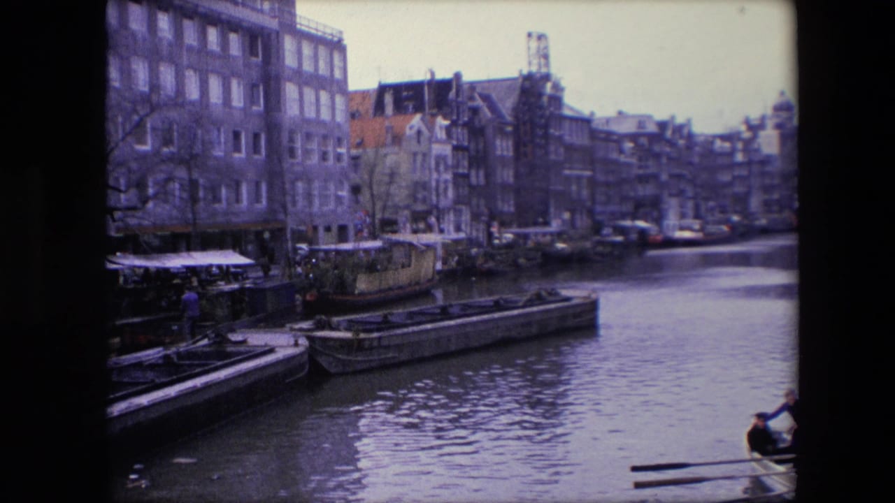 1967 Amsterdam canal view with houseboats, barges, older waterfront buildings, calm water, and two people partly entering the lower right edge of the frame.