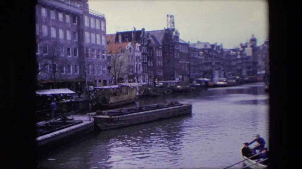 1967 Amsterdam home-movie frame showing tall canal-side buildings, barges, water reflections, and a small rowboat with people near the lower right edge.