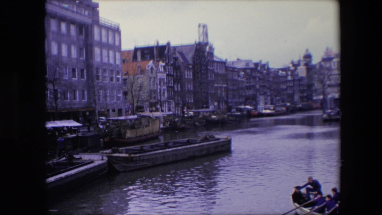 1967 Amsterdam canal view with dense waterfront buildings, moored barges, calm water, and a rowboat carrying visible passengers near the frame edge.