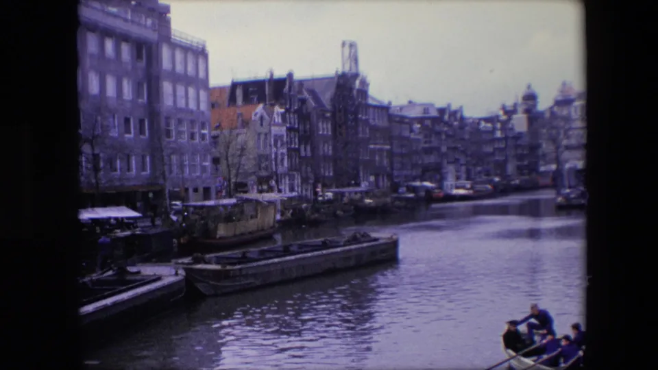 1967 Amsterdam canal view with dense waterfront buildings, moored barges, calm water, and a rowboat carrying visible passengers near the frame edge.