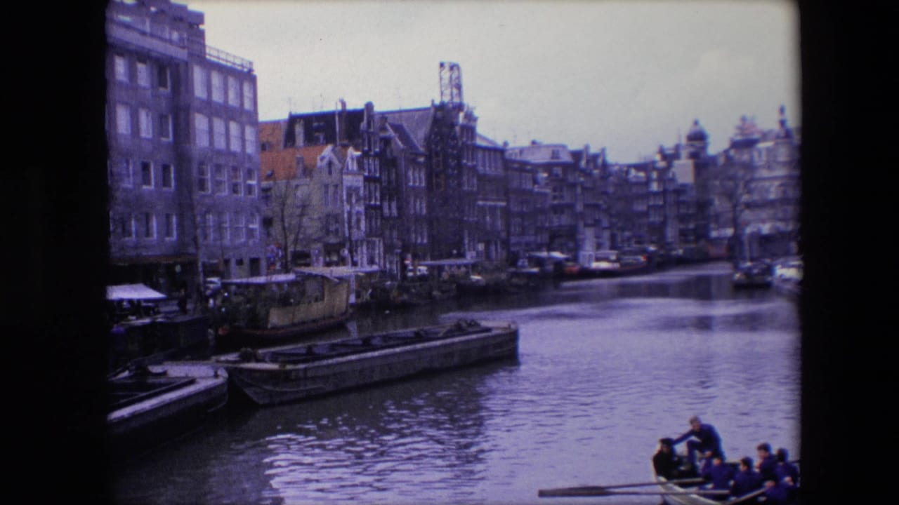 1967 Amsterdam canal frame showing narrow buildings along the water, stationary barges, and a small passenger rowboat partly visible at the bottom.