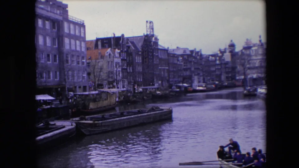 1967 Amsterdam canal frame showing narrow buildings along the water, stationary barges, and a small passenger rowboat partly visible at the bottom.