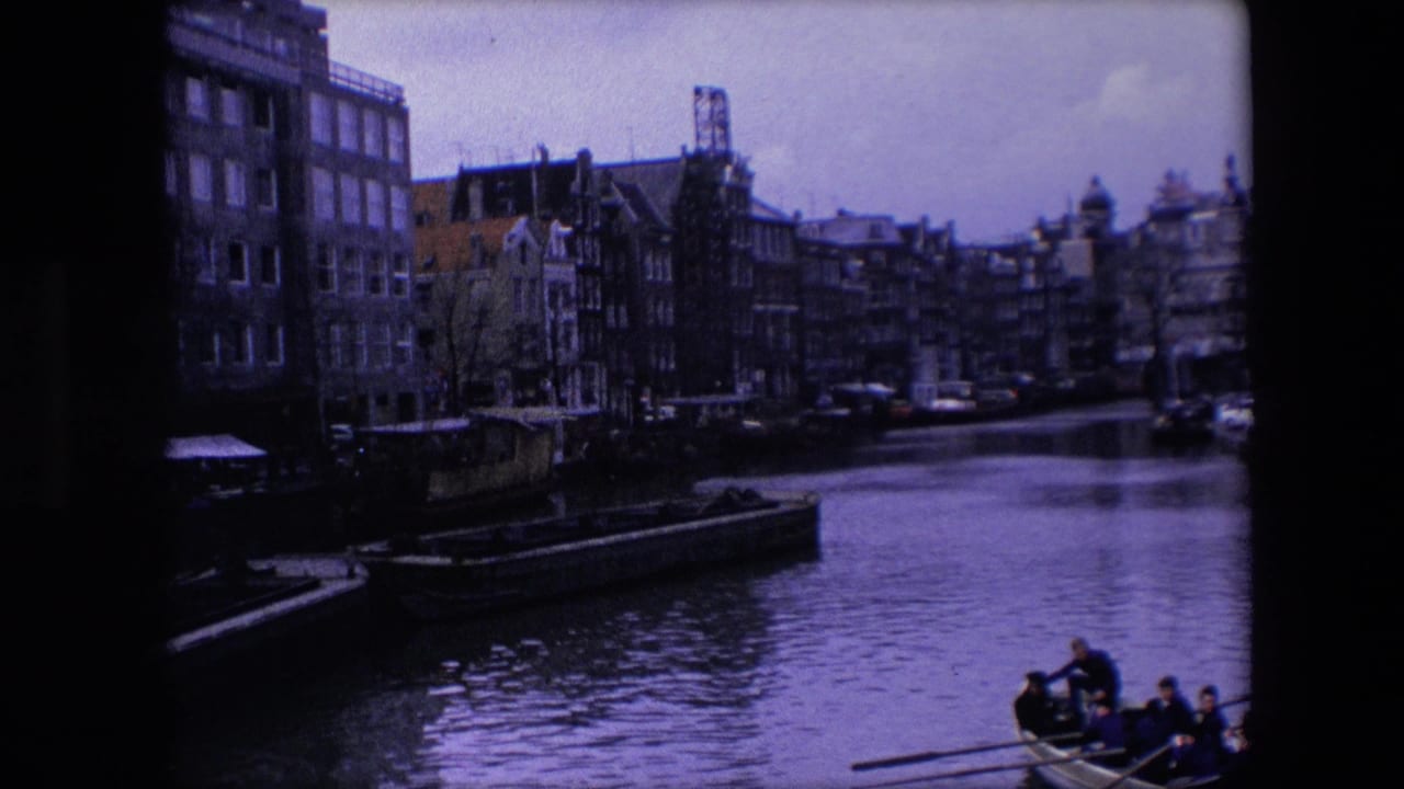 1967 Amsterdam home-movie frame with underexposed canal-side buildings, moored boats, reflective water, and a rowboat with people at the lower right.