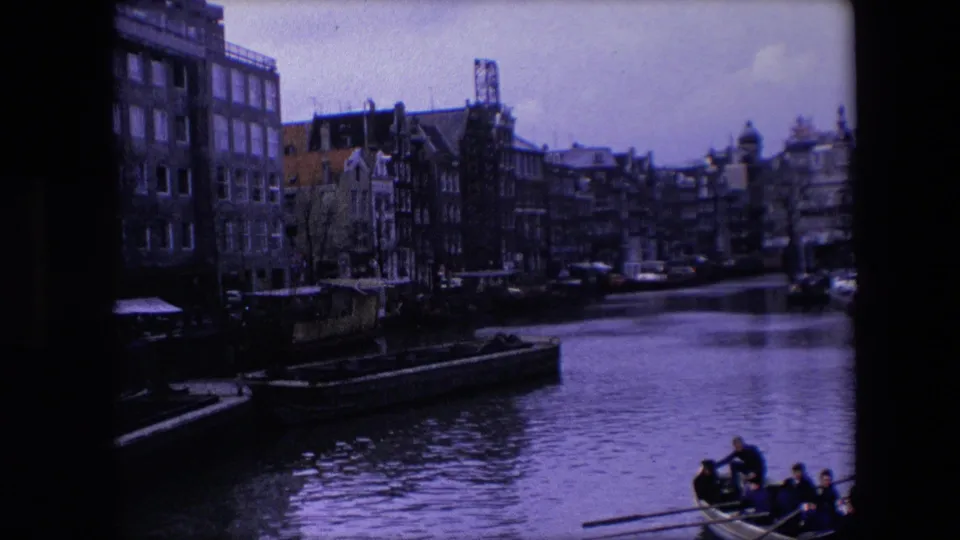 1967 Amsterdam home-movie frame with underexposed canal-side buildings, moored boats, reflective water, and a rowboat with people at the lower right.