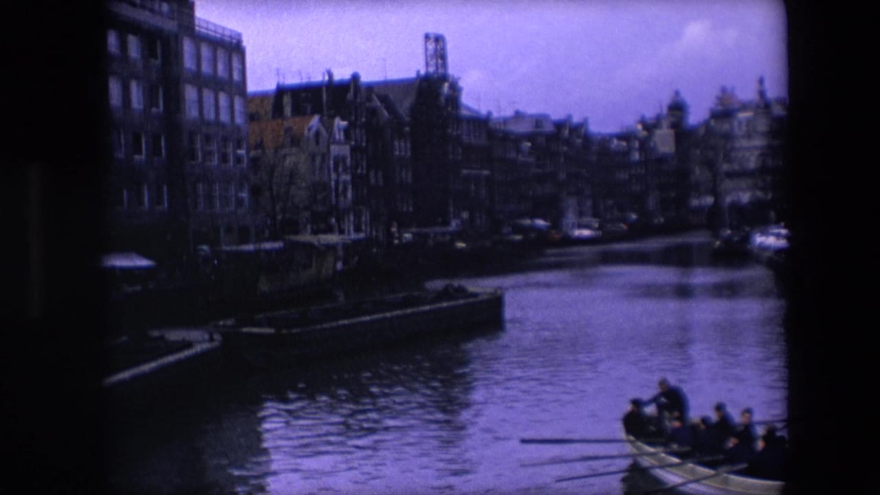 1967 Amsterdam 8mm frame showing a canal bordered by dark brick townhouses, with a rowboat carrying passengers near the lower right edge.