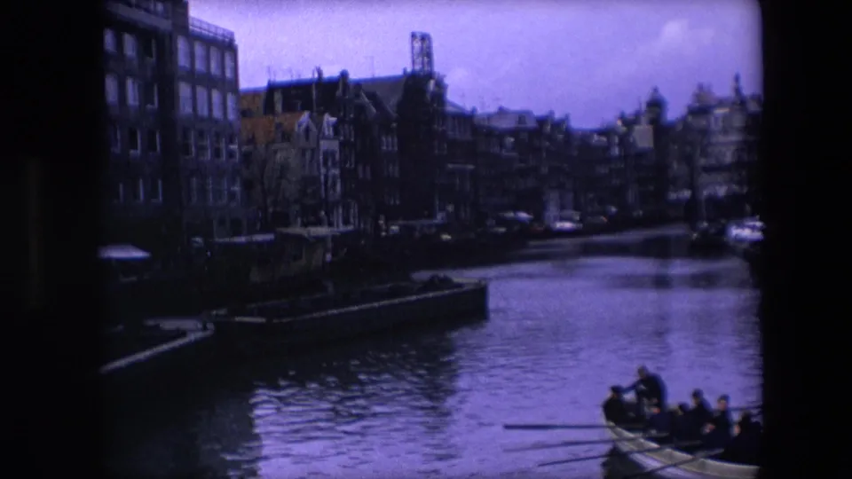 1967 Amsterdam 8mm frame showing a canal bordered by dark brick townhouses, with a rowboat carrying passengers near the lower right edge.