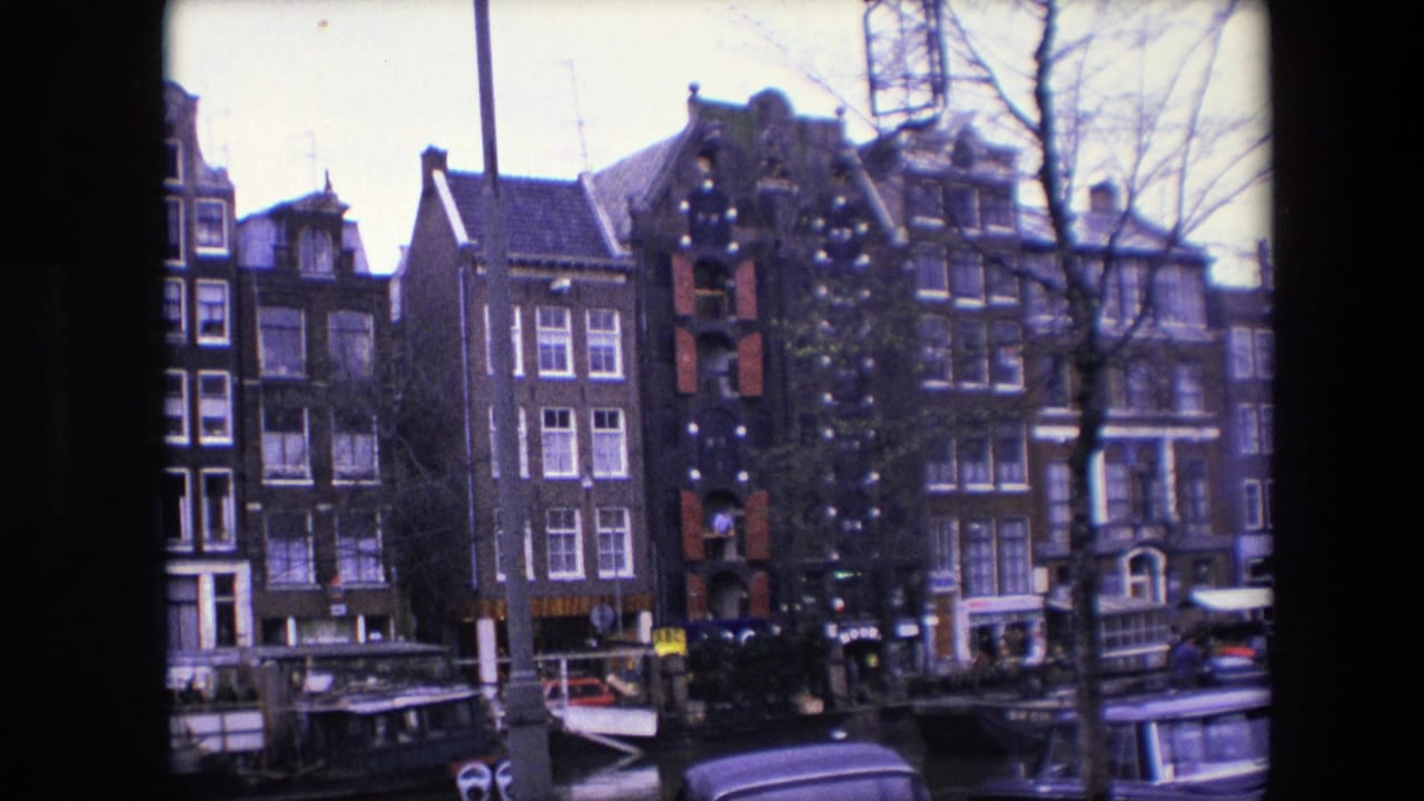 1967 Amsterdam 8mm frame holding on canal-side brick townhouses, bare trees, moored boats, and the roofline of a nearby vehicle.