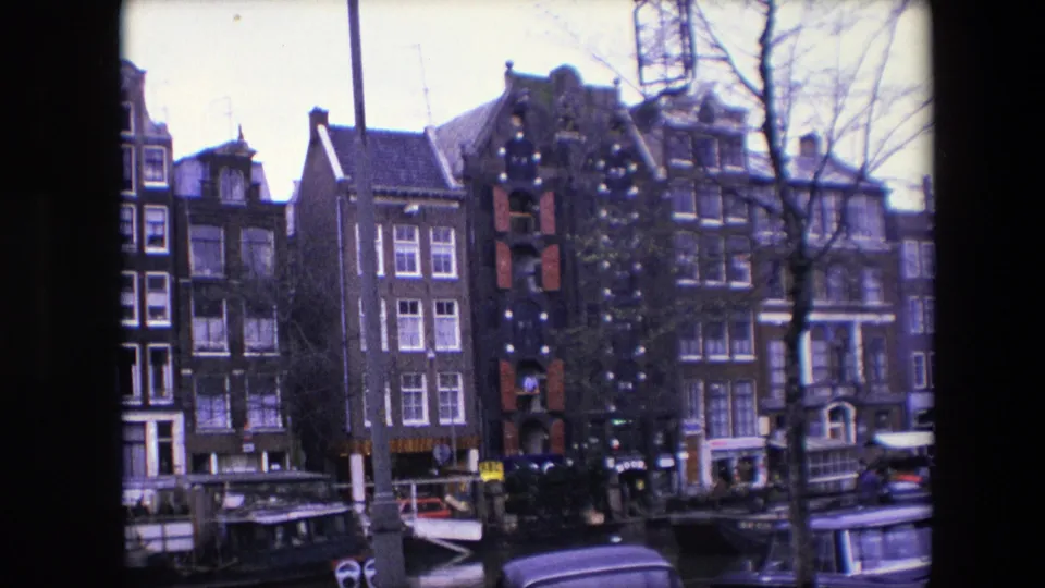 1967 Amsterdam 8mm frame holding on canal-side brick townhouses, bare trees, moored boats, and the roofline of a nearby vehicle.
