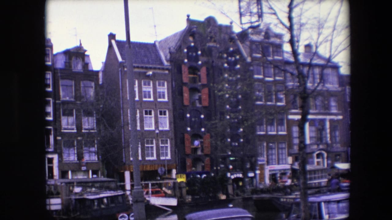 1967 home-movie view of Amsterdam canal houses, including a brick facade with red shutters and exterior decorations, moored boats, bare trees, and small figures near the street.