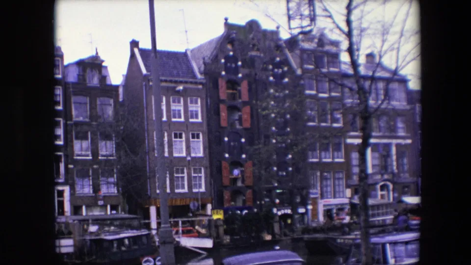 1967 home-movie view of Amsterdam canal houses, including a brick facade with red shutters and exterior decorations, moored boats, bare trees, and small figures near the street.