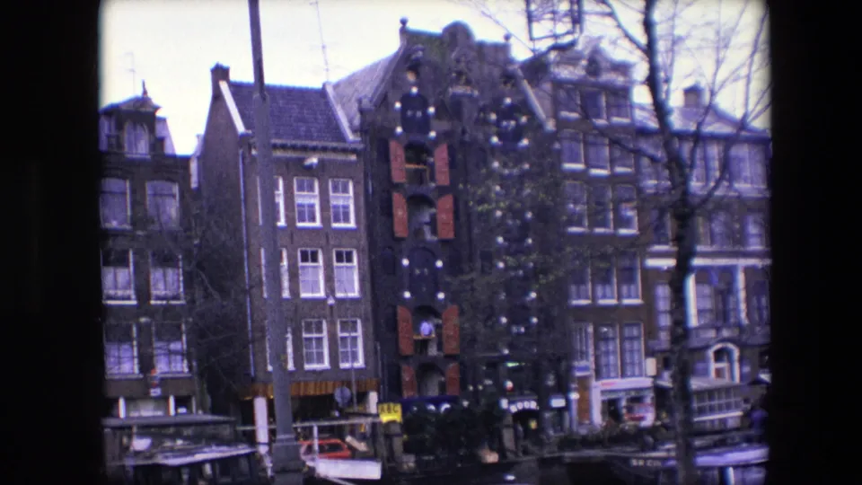 Amsterdam canal-side buildings in 1967, seen from across the water with a decorated central facade, red shutters, moored boats, bare branches, and indistinct people near the lower edge.