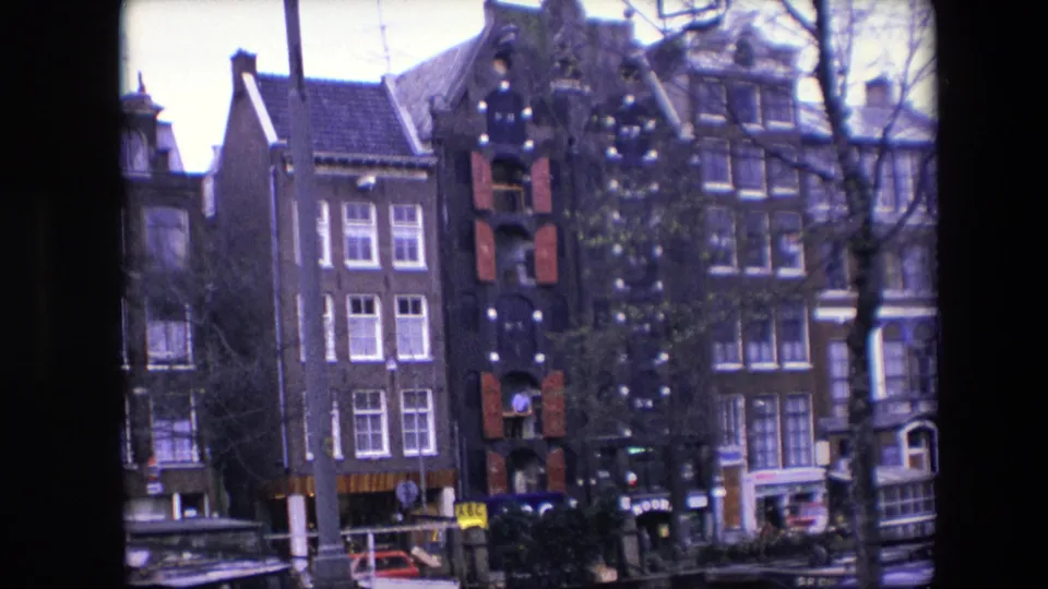 Closer 1967 view of Amsterdam brick canal houses with red shutters, a decorated exterior, bare trees crossing the frame, moored boats, and small street-level figures.