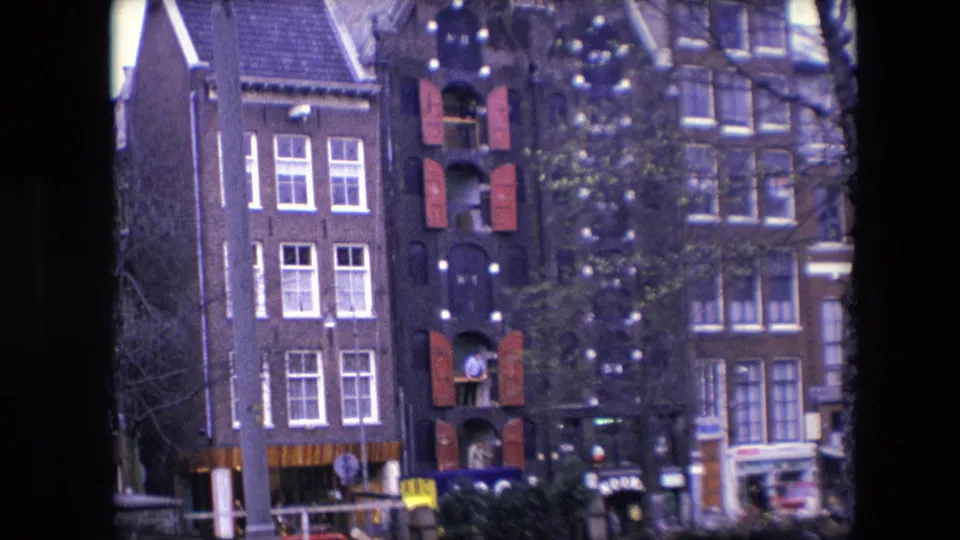 Final frame of the 1967 Amsterdam canal view, centered on a decorated brick building with red shutters, adjacent historic facades, boats below, bare tree branches, and indistinct people or signage at street level.