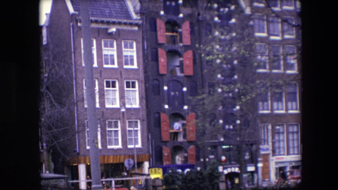 1967 Amsterdam home-movie frame showing a narrow dark brick canal house with red shutters, white-trimmed windows, storefronts, trees, and a man at an upper window.