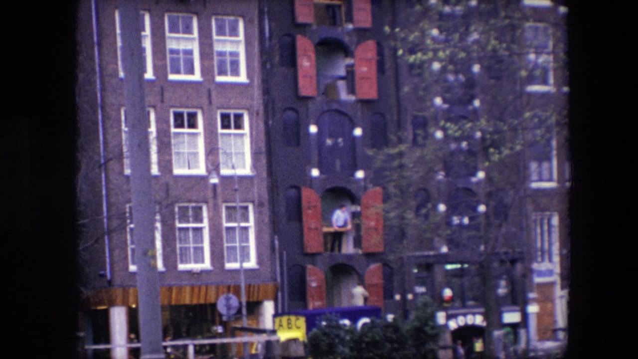Canal house facade with red shutters, street trees, and a man standing in an upper window opening.