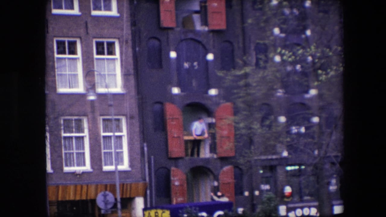 Closer 1967 Amsterdam home-movie view of the dark brick canal house, showing the man at the shuttered upper window with street-level awnings and signs below.