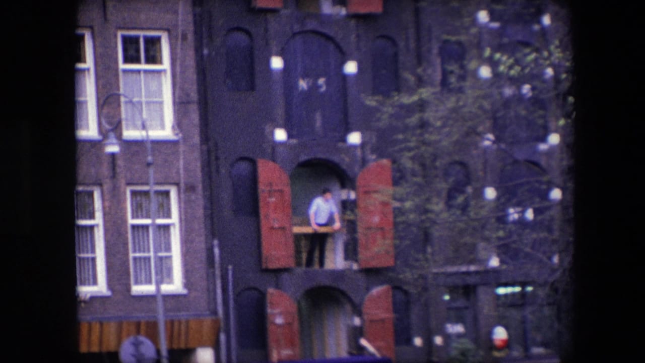 1967 Amsterdam frame showing the shuttered canal-house facade from street level, with a man at an upper opening, white windows, leafy branches, and storefront signage below.