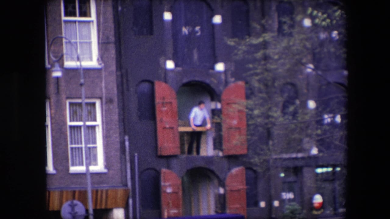 1967 Amsterdam home-movie frame showing an adult man at a second-story opening or small balcony with red shutters on a dark building facade.