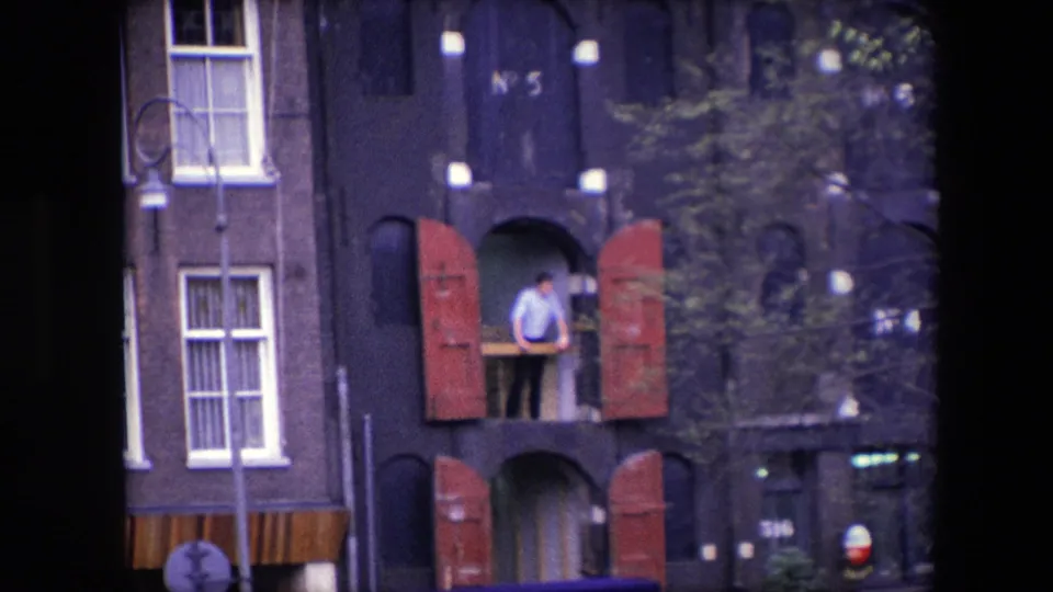 1967 Amsterdam home-movie frame showing an adult man at a second-story opening or small balcony with red shutters on a dark building facade.