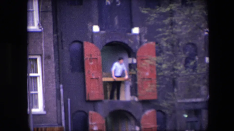 1967 Amsterdam frame with a man at an upper-level opening, red shutters swung outward, and a dark masonry facade partly obscured by nearby foliage.
