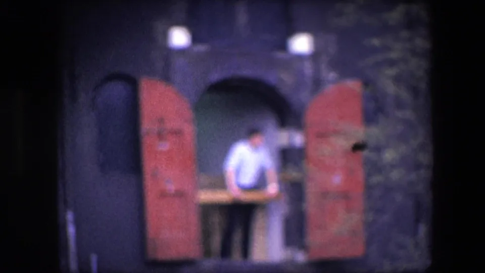 Soft 8mm 1967 Amsterdam close frame showing an adult man at a second-story opening between red shutters, with the dark facade and white window edge nearby.