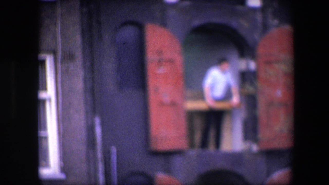 Blurred 1967 Amsterdam home-movie frame with an adult man standing at an upper opening, red shutters on either side, and a dark building facade around him.