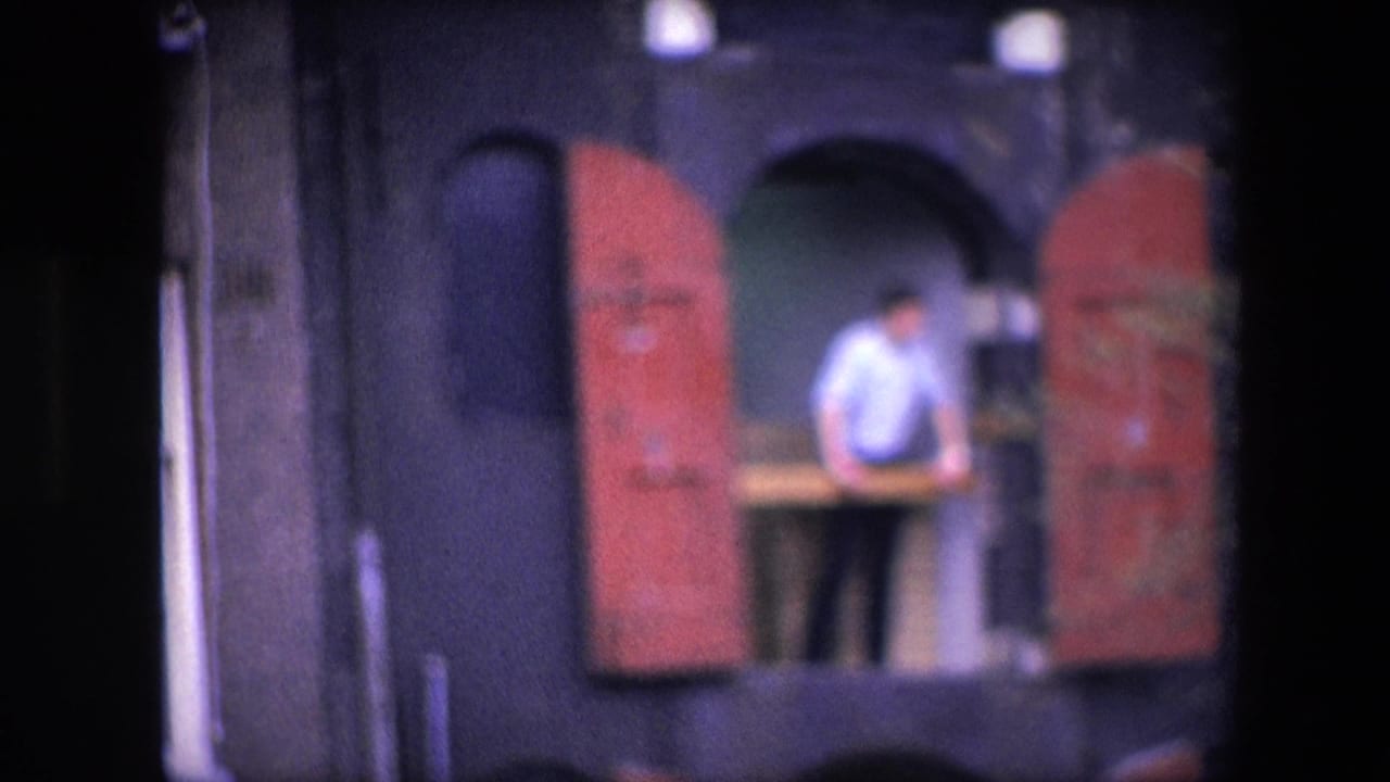 Distant 1967 home-movie frame of a man in a light shirt at an arched balcony opening framed by red shutters, in footage identified as Amsterdam.