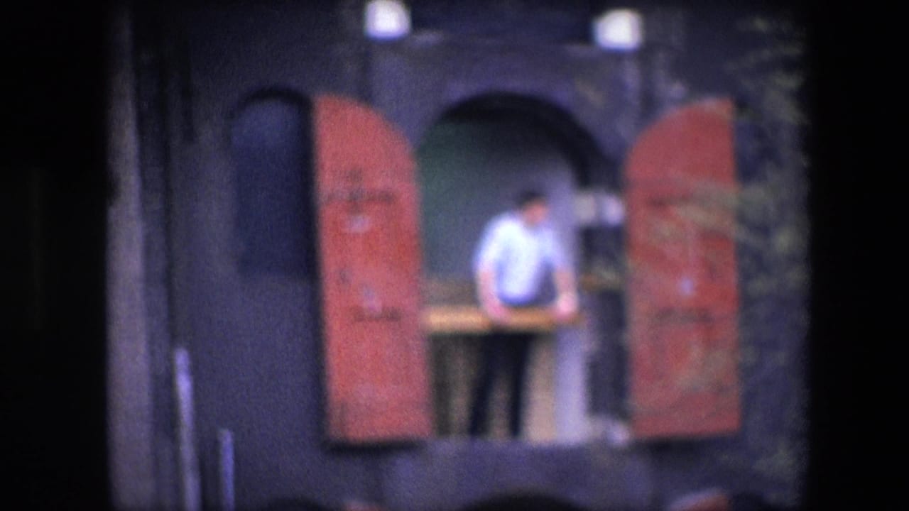 Soft 8mm frame showing the man leaning or standing near the balcony rail, centered between red shutters on a dark facade in the Amsterdam-identified clip.